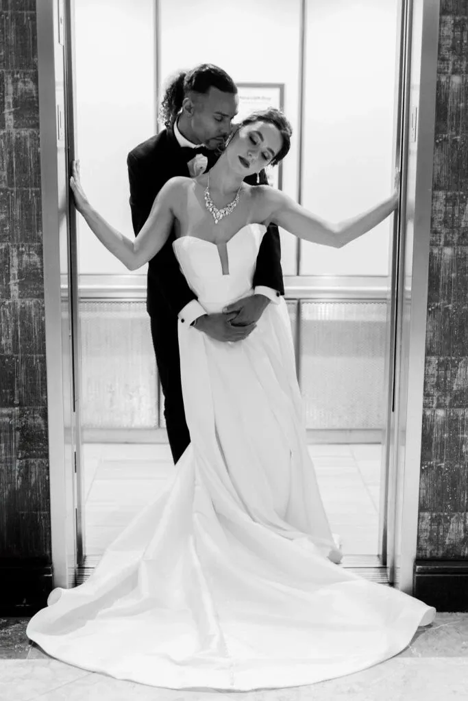 In this black and white image by Braulio Rocha Photography, a couple poses inside an elegant elevator, embodying a romantic and sophisticated wedding scene. The bride is wearing a strapless, flowing wedding gown with a sweetheart neckline, complemented by a statement necklace, while the groom stands behind her in a classic black tuxedo. The couple's intimate pose, with the bride leaning against the elevator doors and the groom gently embracing her, captures their connection beautifully. This image exemplifies the elegance and artistry characterized by Braulio Rocha Photography's wedding photography in Canada.