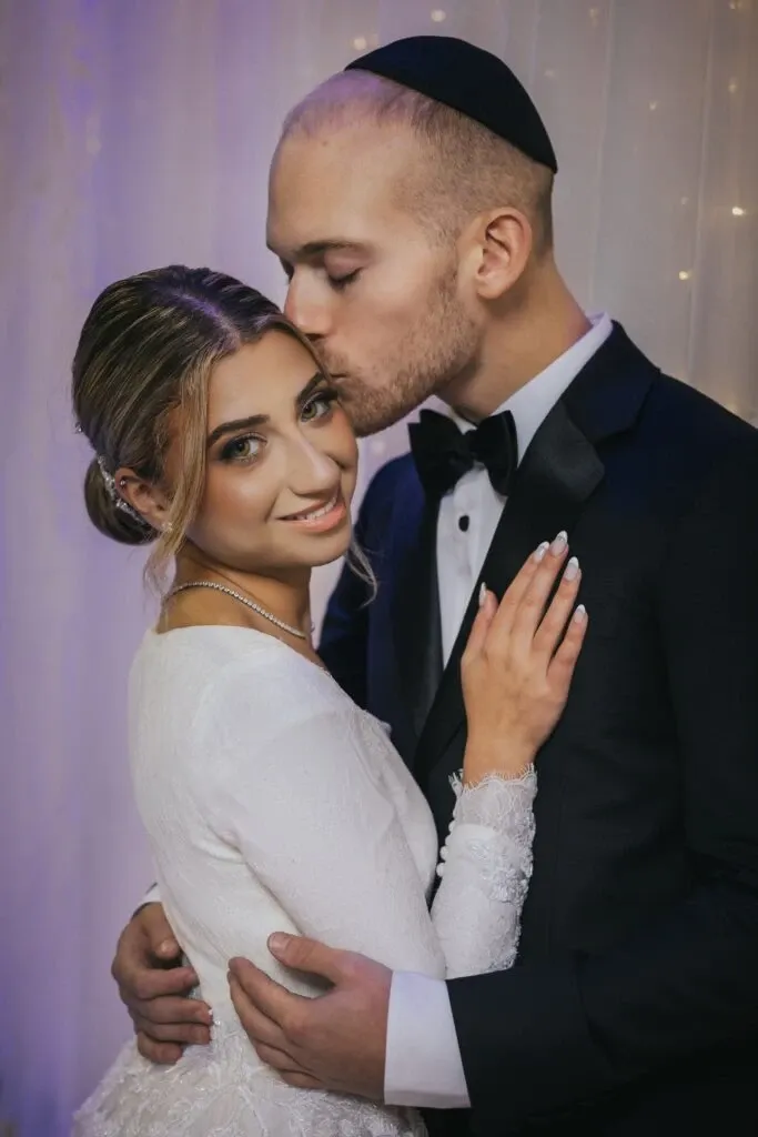 This image by Braulio Rocha Photography, featured in the wedding photography category, captures a tender moment between a newlywed couple. The groom, dressed in a classic black tuxedo and wearing a traditional kippah, gently kisses the bride's forehead. The bride, in an elegant white lace gown, smiles warmly at the camera. They are embraced against a softly lit backdrop, creating a romantic and intimate atmosphere, ideal for showcasing natural, emotional wedding photography.