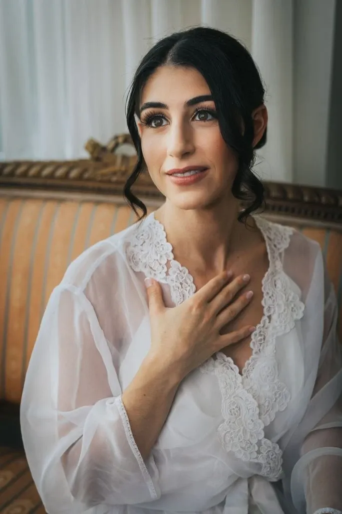 A bride is seated on an elegant, vintage couch adorned with soft, striped fabric. She is wearing a delicate, white lace bridal robe, showcasing intricate floral patterns along the edges. Her dark hair is styled in loose, romantic curls. Her expression is one of joyful anticipation, with her hand gently placed on her chest. The atmosphere is intimate and serene, with soft natural light filtering through white curtains. Captured by Braulio Rocha Photography, this image exemplifies the category of Wedding Photography, highlighting a candid moment of reflection before the ceremony.