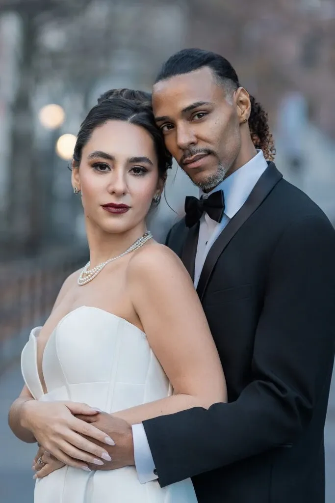 In this elegant wedding photography image by Braulio Rocha Photography, a newlywed couple is captured in a serene outdoor setting. The bride, dressed in a strapless white wedding gown with a subtle sweetheart neckline, is adorned with a delicate necklace and matching earrings. Her hair is styled in a classic updo, adding to the sophisticated look. The groom stands beside her, wearing a sleek black tuxedo with a black bow tie. He has one arm around her waist, and they both gaze softly at the camera, exuding love and happiness. The soft, blurred background suggests a romantic urban environment, enhancing the intimate moment captured in this wedding photography piece.