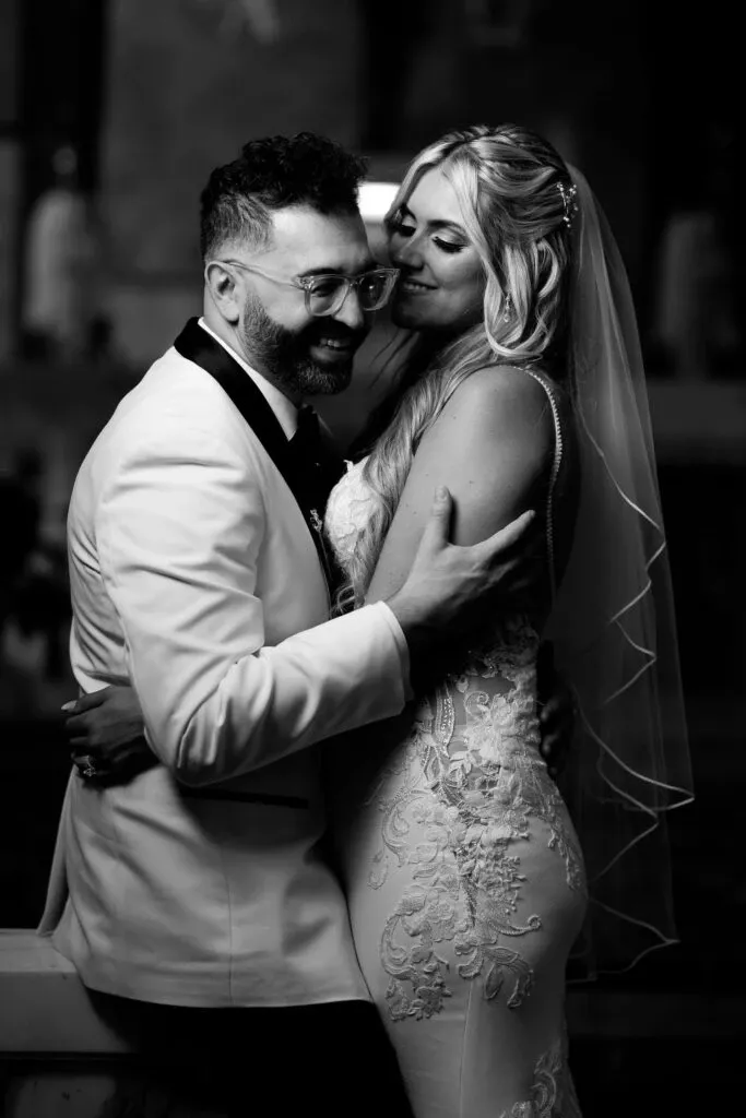 A bride and groom share an intimate moment, captured by Braulio Rocha Photography in the wedding photography category. The bride, with long, flowing hair and a delicate veil, wears an intricately embroidered lace gown, while the groom, wearing glasses and a stylish white tuxedo with a black lapel, embraces her lovingly. The black and white image highlights the couple's closeness and joy on their special day, representing the elegance and artistry typical of wedding photography by Braulio Rocha Photography, featured on the Mariage Elegant, Canada website.
