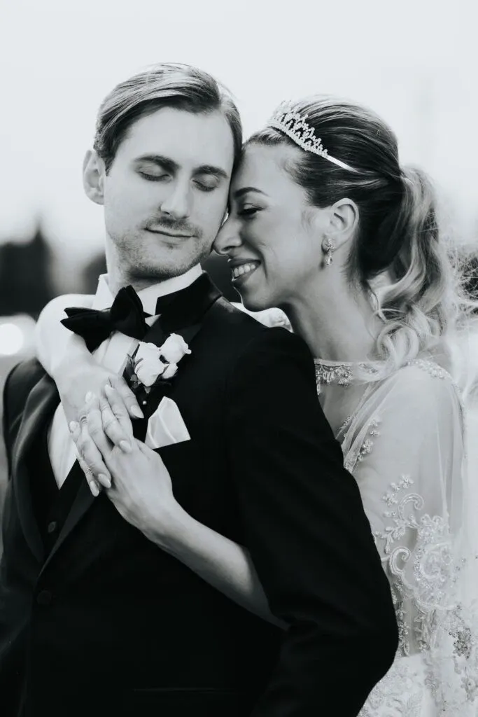This black and white photograph by Braulio Rocha Photography, available under the Wedding Photography category on Mariage Elegant, Canada, beautifully captures an intimate moment between a bride and groom. The groom is dressed in a classic black tuxedo with a crisp white shirt and a bow tie, complemented by a boutonnière. The bride, adorned with an elegant tiara, is wearing a detailed lace wedding dress and gently embraces the groom, showcasing her delicate wedding ring. This touching image reflects the couple's joy and connection on their special day, a perfect keepsake captured by a skilled wedding photographer.