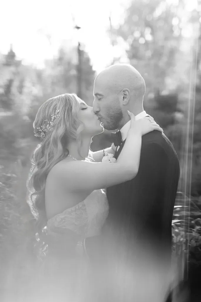 In this elegant image captured by Braulio Rocha Photography, a bride and groom share a tender kiss in an outdoor setting, highlighting the wedding photography category. The bride wears a strapless gown adorned with intricate lace details and a delicate floral headpiece in her flowing hair. The groom, dressed in a classic tuxedo, holds the bride close. Soft natural light filters through the surrounding trees, creating a dreamy and romantic atmosphere, ideal for capturing memorable wedding moments.