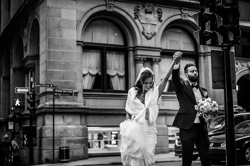 In the image captured by Inhouse Studios Photography, a bride and groom joyously walk hand-in-hand on a city street, exemplifying the charm of urban wedding photography. The bride, in an elegant wedding gown with a lace veil, playfully lifts her dress as she smiles. The groom, dressed in a classic tuxedo, holds a bouquet of flowers while confidently leading the way. The backdrop features a historic building with ornate stonework and large arched windows, adding a timeless and sophisticated ambiance to their wedding day. This candid moment beautifully illustrates the essence of wedding photography by Inhouse Studios Photography, highlighting the joy and romance of the occasion.