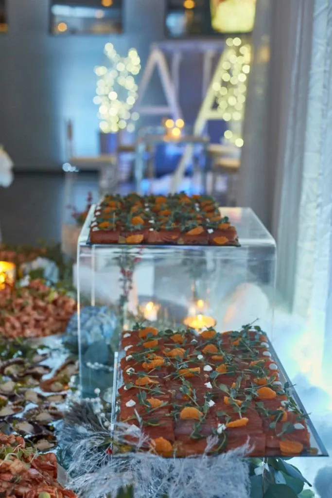 The image features an elegantly arranged wedding reception setup at Plaza Du Parc, a premier wedding venue in Canada. In the foreground, there is a clear display case showcasing delicately crafted hors d'oeuvres garnished with fresh herbs and small slices of fruit, highlighting the venue's attention to detail in catering. The background is softly illuminated with warm, ambient lighting and decorative twinkling lights, creating a romantic atmosphere perfect for wedding celebrations. This setting exemplifies the sophisticated and stylish event spaces offered by Plaza Du Parc for memorable wedding ceremonies and receptions.