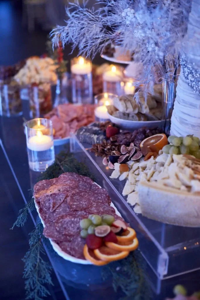 The image showcases a beautifully arranged charcuterie table at Plaza Du Parc, a wedding venue in the Mariage Elegant vendor listings. The table is adorned with a variety of cured meats, including salami arranged alongside fresh grapes and orange slices. A selection of cheeses is artfully displayed, complemented by nuts and dried fruits. Soft candlelight adds a romantic ambiance, enhancing the elegant presentation with feathery floral decorations. This setting highlights the venue's attention to detail and sophistication, ideal for a memorable wedding celebration.
