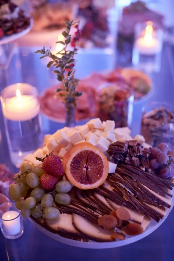 In this image from the wedding venue Plaza Du Parc, categorized under Wedding Venues on Mariage Elegant, Canada, there is an elegantly arranged charcuterie board that serves as a centerpiece. The board features an array of fresh fruits, including strawberries, grapes, and a sliced blood orange. It is complemented by cubes of cheese and an assortment of nuts, all presented on a glass surface. Soft candlelight in the background adds a warm ambiance, enhancing the sophisticated presentation perfect for a wedding reception.