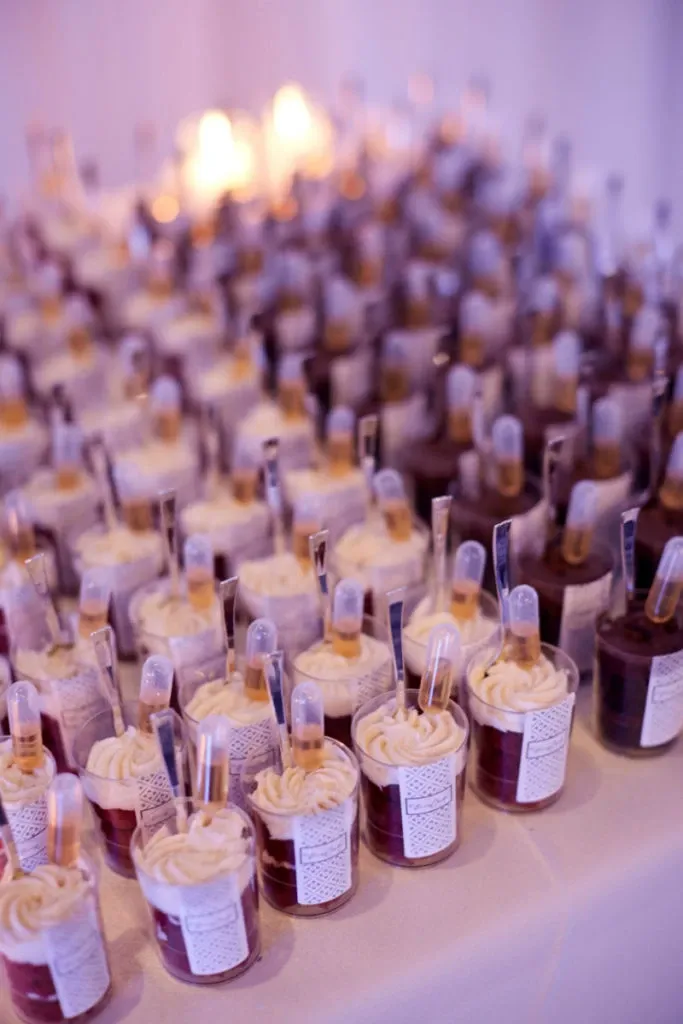 In the image, a beautifully arranged table is filled with individual dessert cups, each topped with swirls of whipped cream and a small pipette filled with sauce for added flavor. These elegant treats are part of the catering display at Plaza Du Parc, a featured wedding venue in Canada on Mariage Elegant. The soft lighting and artful arrangement suggest a sophisticated and inviting atmosphere, typical of the wedding venues category, highlighting Plaza Du Parc's attention to detail and commitment to a luxurious experience.