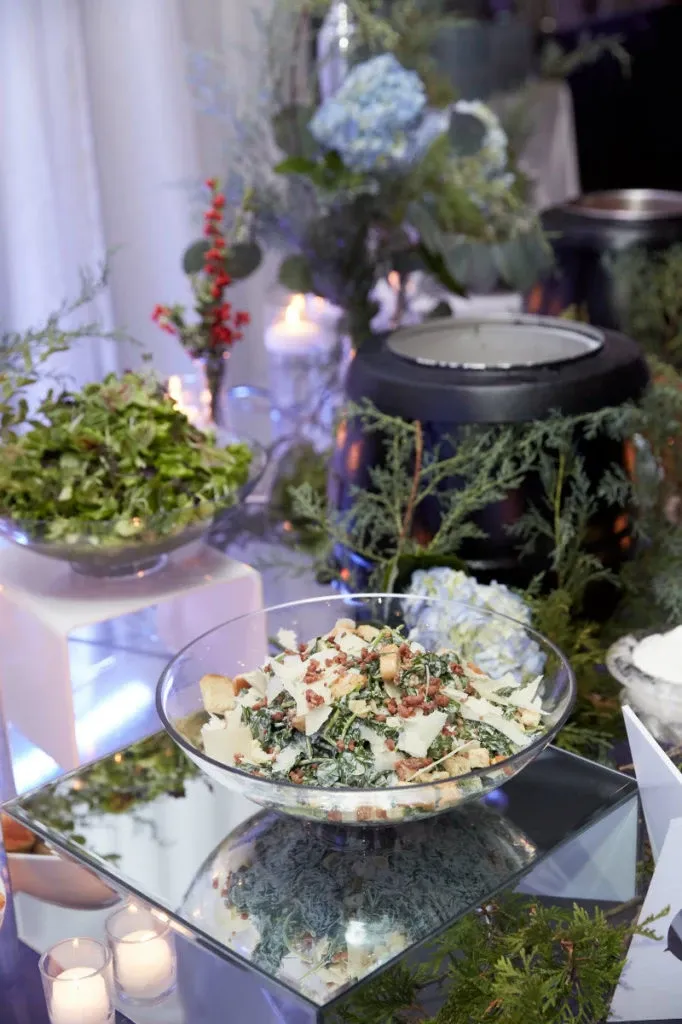 The image shows an elegant buffet setup at Plaza Du Parc, a premier wedding venue in Canada, listed on Mariage Elegant. The table features a visually appealing salad in a glass bowl atop a reflective mirror surface, garnished with sliced almonds and shaved cheese. Surrounding the salad are vibrant floral arrangements, including blue hydrangeas and red berries, enhancing the romantic ambiance. Two white candles in glass holders provide soft lighting, while a black pot warmer in the background suggests a variety of food options available for wedding guests. This setup highlights the attention to detail and sophistication Plaza Du Parc offers as a wedding venue.