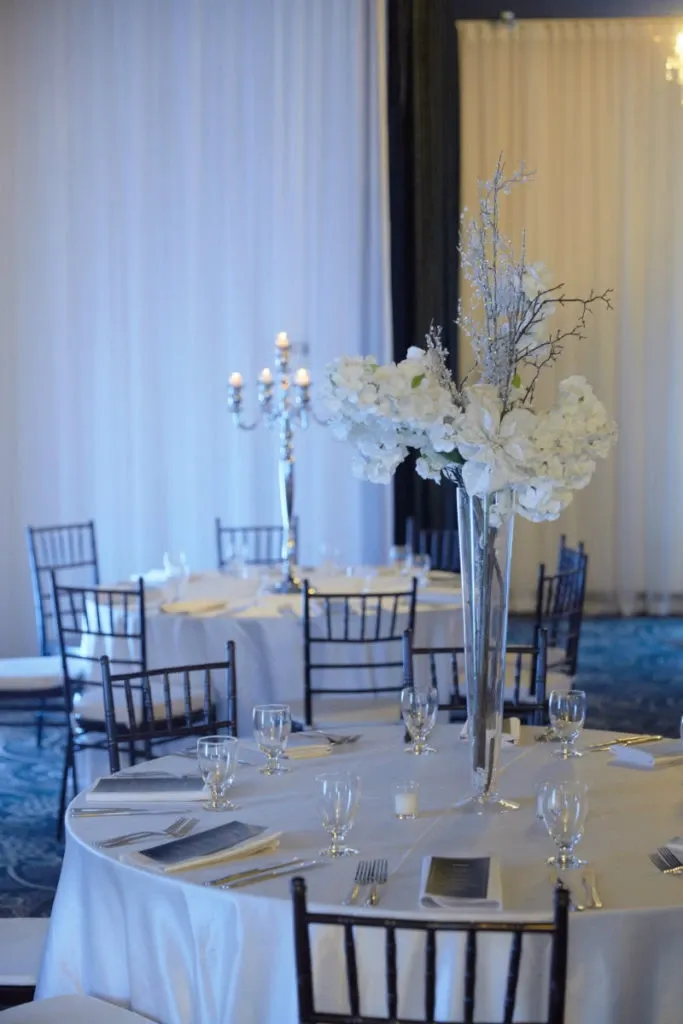 The image features an elegant wedding venue setup at Plaza Du Parc, highlighting its suitability within the Wedding Venues category. The room is adorned with beautifully arranged tables, each draped in crisp white tablecloths and decorated with tall vases containing white floral arrangements. Visitors can notice the silver candelabras adding a sophisticated touch, casting a warm glow that enhances the romantic ambiance. Black chairs surround the tables, and neatly placed glassware and cutlery complete the refined table settings. The venue's subtle blue and white color scheme and exquisite decor details contribute to an atmosphere ideal for hosting an unforgettable wedding celebration.