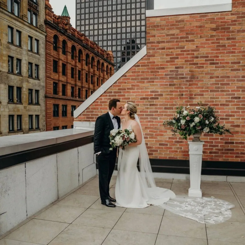 In this image titled 'Corner Collection' under the category of Wedding Venues on Mariage Elegant, Canada, a bride and groom share a tender kiss on a rooftop terrace. The bride is dressed in a flowing white gown with a long train, complemented by a delicate veil. She holds a lush bouquet of white and soft pink flowers. The groom is in a classic black suit. Beside them stands a large floral arrangement on a white pedestal. The background features charming brick buildings, creating an elegant urban atmosphere for this intimate wedding moment.
