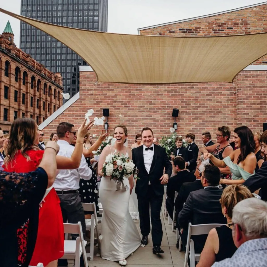 In this image, a joyous couple walks down the aisle at their wedding ceremony, held at Corner Collection, a featured wedding venue on Mariage Elegant, Canada. The bride, in an elegant white gown, holds a bouquet of pastel flowers, while the groom is dressed in a classic black tuxedo. The setting is an outdoor rooftop venue with a charming mix of historic brick architecture and modern urban views. Guests, dressed in formal attire, are seated and cheering, creating a lively and celebratory atmosphere. Decor includes white seating and a beige shade sail overhead, adding to the venue's sophisticated and stylish ambiance.