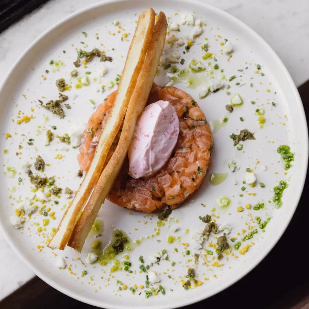 The image showcases an elegant dish served at Corner Collection, a distinguished wedding venue listed on Mariage Elegant, Canada. The plate features a finely diced salmon tartare accompanied by a dollop of cream. Garnished with vibrant green herbs and a sprinkle of seasoning, the presentation is enhanced by two crisp breadsticks artfully placed on top. This gourmet dish epitomizes the culinary excellence that Corner Collection offers as part of their wedding venue services, ensuring a memorable dining experience for wedding guests.