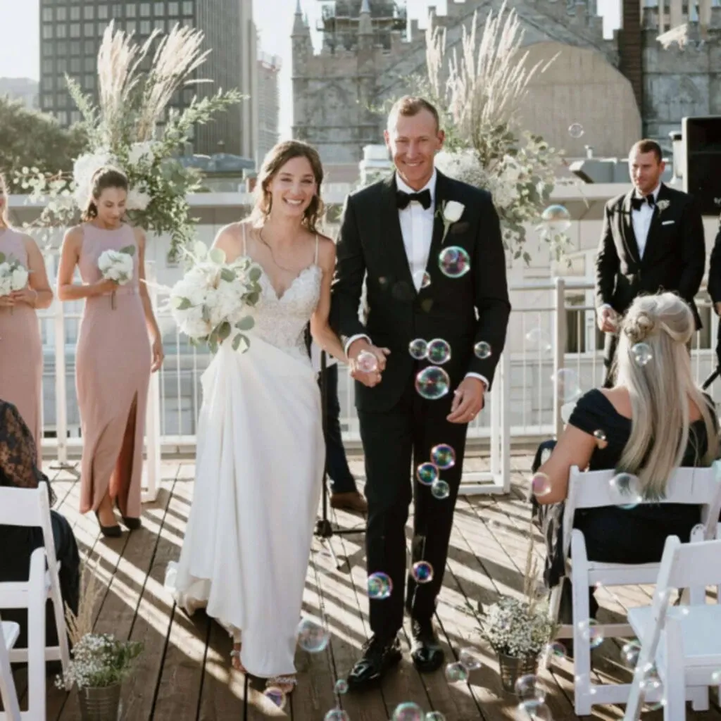 The image shows a joyful couple at Corner Collection, a premier wedding venue, part of the Wedding Venues category for Mariage Elegant in Canada. The bride, in an elegant white gown with lace detailing, and the groom, in a classic black tuxedo, are walking hand-in-hand down the aisle. They are surrounded by guests seated on white chairs on a wooden deck adorned with delicate floral arrangements and tall pampas grass. A whimsical touch is added by floating bubbles in the air. The setting is outdoors, with a cityscape visible in the background, creating a perfect blend of urban charm and romantic ambiance for a wedding ceremony.
