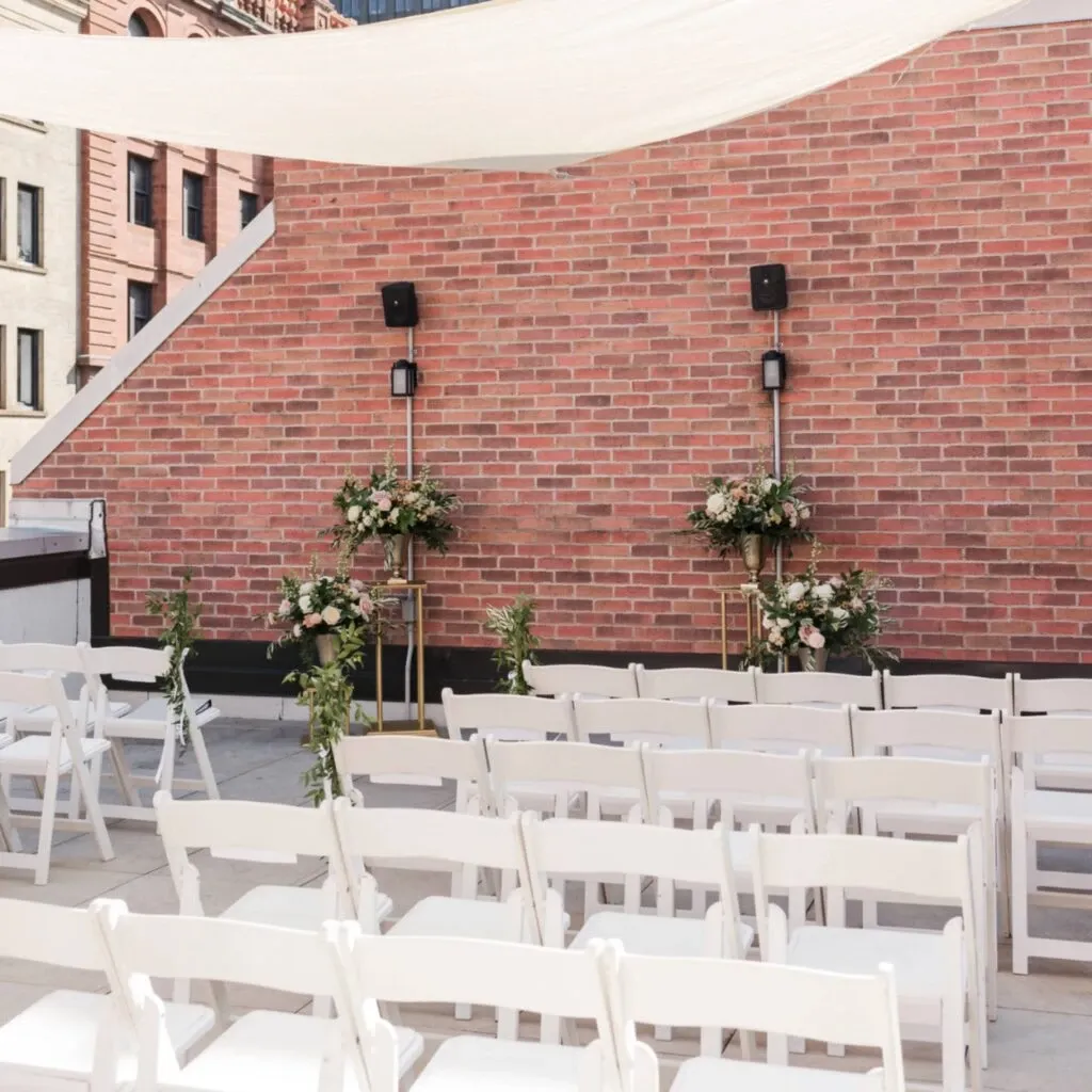 The image shows an elegant rooftop wedding venue by Corner Collection, categorized under Wedding Venues. Rows of white chairs are neatly arranged facing a backdrop of a red brick wall. Two large floral arrangements filled with white and soft pink flowers, accented with green leaves, are elegantly placed on gold stands against the wall. Above the seating area, a sheer white canopy provides shade, creating a bright and inviting atmosphere. This setup highlights the chic and intimate ambiance perfect for a urban rooftop wedding in Canada.