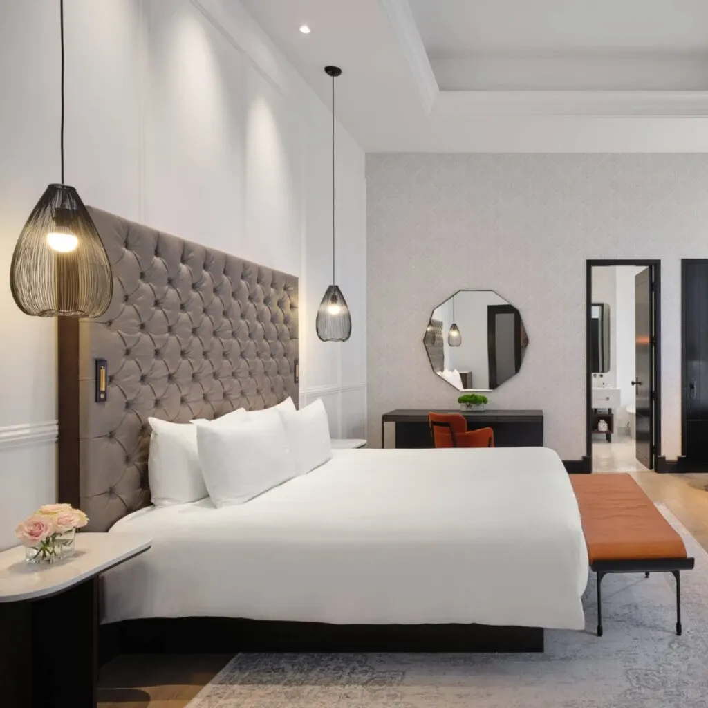 This image features an elegantly designed room at Corner Collection, a wedding venue listed under the Wedding Venues category on Mariage Elegant, Canada. The sophisticated bedroom showcases a plush, tufted headboard against a soft white wall, complemented by modern pendant lights hanging on either side of the bed. A round mirror decorates the wall above a sleek wooden desk, with a small green plant adding a touch of nature. A cozy bench rests at the foot of the bed, while vibrant pink and white roses adorn a bedside table, creating a romantic and inviting atmosphere ideal for newlyweds.