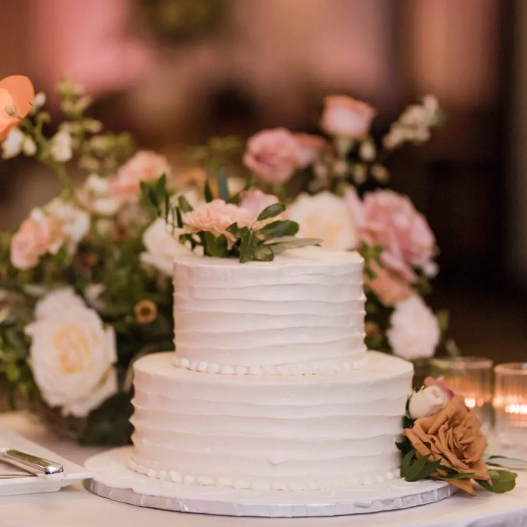 This image showcases a beautifully decorated wedding venue from Corner Collection, featured in the Wedding Venues category. The focal point is an elegantly crafted two-tiered white wedding cake adorned with delicate pink and peach floral accents. The cake is set against a backdrop of a lush floral arrangement featuring roses in complementary pastel hues, providing a romantic and sophisticated atmosphere. Soft candlelight adds a warm glow, enhancing the intimate setting ideal for a wedding celebration.