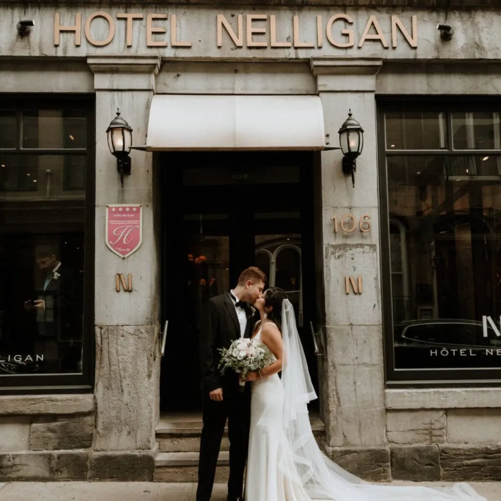 A newlywed couple is sharing a romantic moment in front of Hotel Nelligan, a sophisticated venue included in Corner Collection's offerings for wedding venues. The bride is wearing an elegant white gown with a long veil, holding a beautiful bouquet of flowers. The groom is dressed in a classic black tuxedo. The hotel's historic stone facade and stylish entrance enhance the intimate ambiance, perfect for weddings planned through Mariage Elegant in Canada.