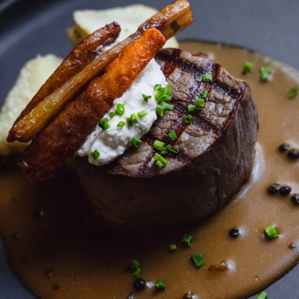 This image showcases a beautifully presented gourmet dish at Corner Collection, a premium wedding venue listed under Mariage Elegant in Canada. The plate features a perfectly cooked filet mignon topped with a dollop of creamy cheese and garnished with vibrant green chives. Accompanying the steak are crispy, golden brown carrot batons elegantly arranged on top. The dish is artistically placed on a bed of rich, flavorful sauce, dotted with whole peppercorns, adding an exquisite touch to the culinary presentation. This elegant meal highlights the sophisticated dining experience offered by Corner Collection, enhancing the appeal of your wedding celebration.