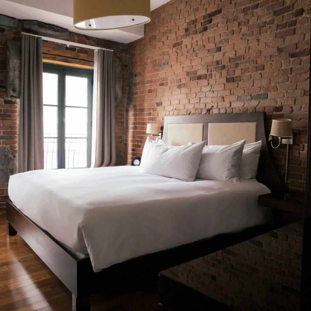 The image showcases an elegant room at Corner Collection, a wedding venue listed under Mariage Elegant, Canada. The room features a large, neatly made bed with crisp white linens, set against a rustic, exposed brick wall. Two modern wall sconces flank the headboard, providing soft lighting. A large window with curtains filters natural light into the space, highlighting the wooden flooring. This venue offers a warm and inviting atmosphere, ideal for intimate wedding ceremonies and bridal preparations.