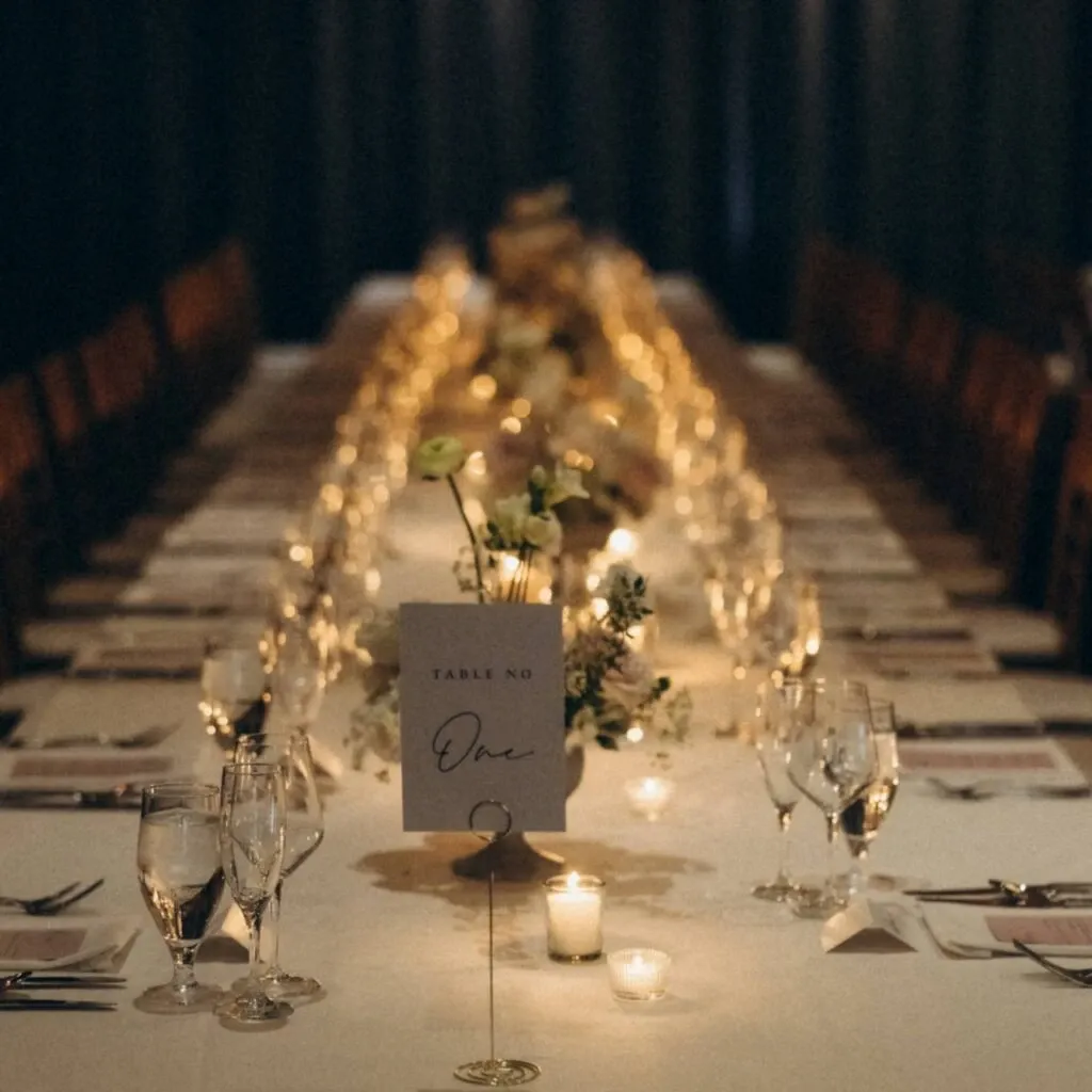 The image showcases an elegant wedding reception table arrangement by Corner Collection, categorized under Wedding Venues. The table is beautifully set with white linens, creating a refined atmosphere. Soft, glowing candlelight adds a warm ambiance, enhancing the sophisticated setting. Delicate floral centerpieces with white and light green flowers adorn the table, while neatly folded napkins and polished glassware complete the arrangement. A table number card labeled 'One' is prominently displayed, reflecting a well-organized event setting suitable for a memorable celebration.