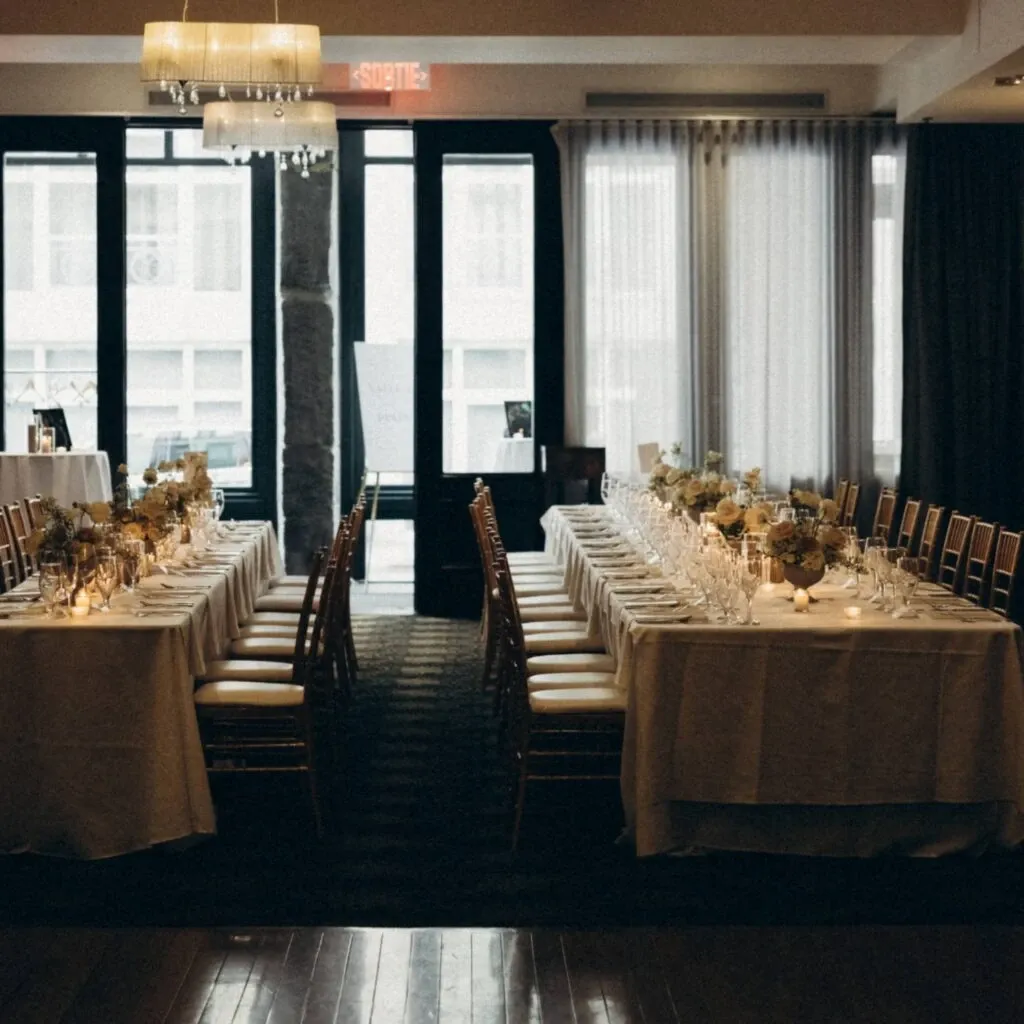 The image features an elegantly set wedding venue by Corner Collection, under the wedding venues category on Mariage Elegant, Canada. The room showcases two long banquet tables adorned with white tablecloths and lined with gold chairs, creating a sophisticated and inviting atmosphere. Each table is beautifully decorated with floral centerpieces and softly lit candles, enhancing the romantic ambiance. Large windows in the background allow natural light to stream in, complemented by a modern chandelier hanging from the ceiling. The venue exudes a chic and intimate setting, perfect for a wedding celebration.