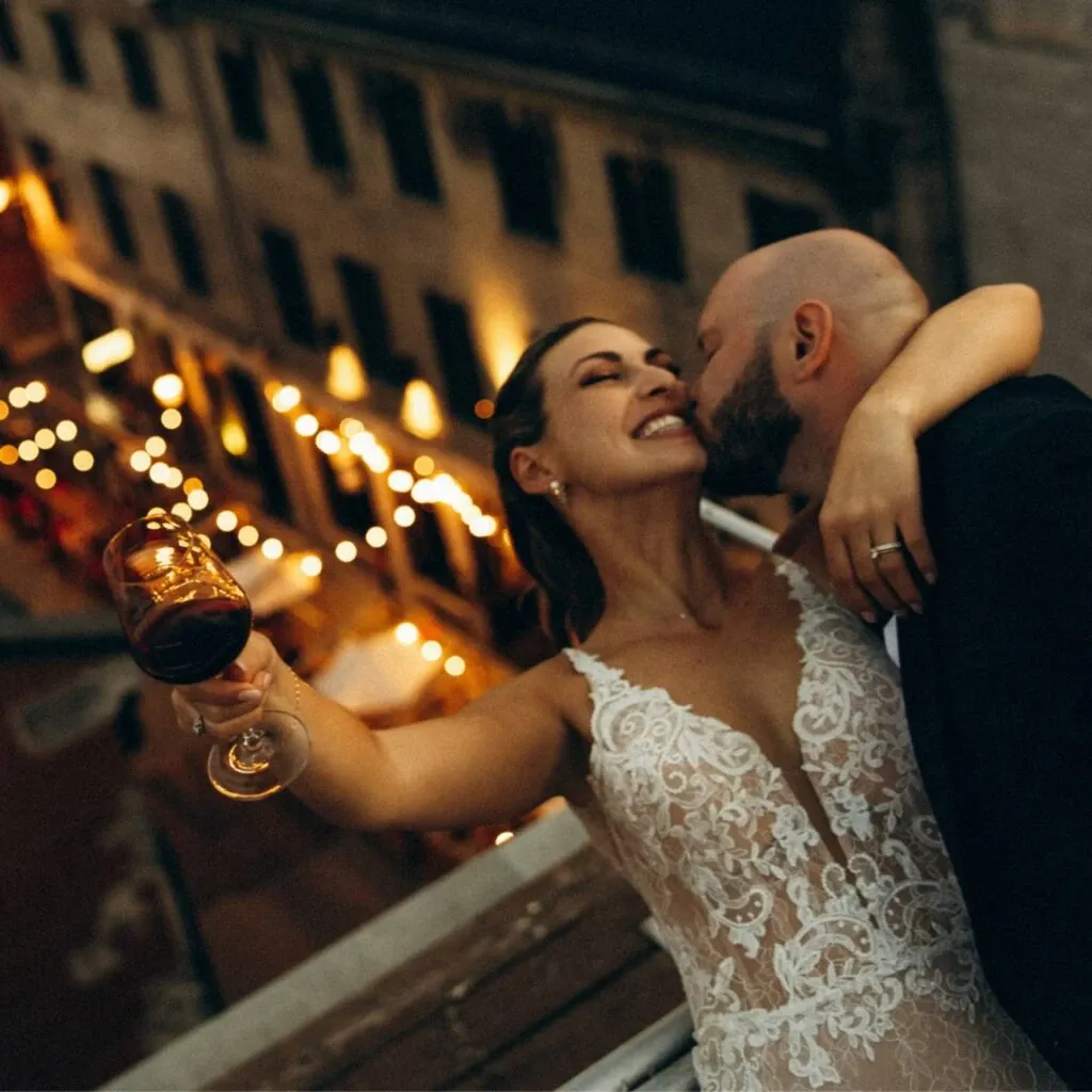 A joyful couple embraces on a balcony at a wedding venue provided by Corner Collection, a top vendor in the Wedding Venues category. The bride, wearing a detailed lace wedding gown, holds a glass of red wine, smiling gleefully as the groom, in formal attire, kisses her cheek. Below, warm string lights illuminate an intimate outdoor setting, capturing the romantic ambiance typical of Corner Collection's picturesque venues in Canada.