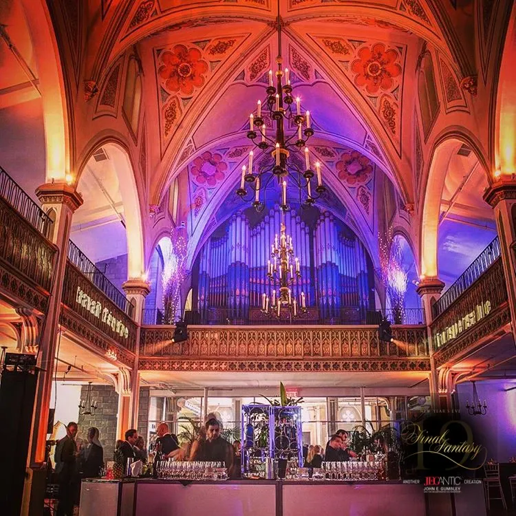 The image showcases the elegant interior of Le Salon Richmond, a wedding venue listed under Mariage Elegant in Canada. The venue features a grand, vaulted ceiling adorned with intricate floral designs and illuminated with soft, ambient lighting. A large, ornate chandelier hangs prominently in the center, casting a warm glow over the space. Beneath the chandelier, a bar area is set up with glasses neatly arranged, and several people are gathered, engaged in conversation, enhancing the lively, celebratory atmosphere. This venue blends historic architectural beauty with modern wedding functionality.