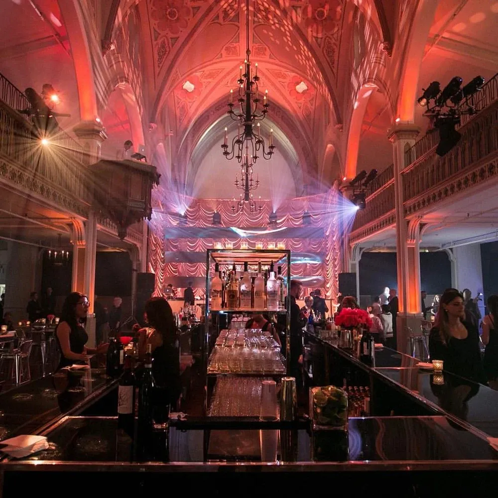 The image showcases an elegant wedding venue by Le Salon Richmond, categorized under Wedding Venues. The scene is set in a grand hall with a high, vaulted ceiling adorned with intricate architectural details. Ambient lighting in warm red hues casts a romantic glow over the space. A central chandelier adds a touch of sophistication. In the foreground, there is a long, polished bar with glassware neatly arranged, surrounded by attendees in formal attire. The background reveals an area prepared for an event, featuring draped curtains and atmospheric lighting, ideal for weddings or receptions.