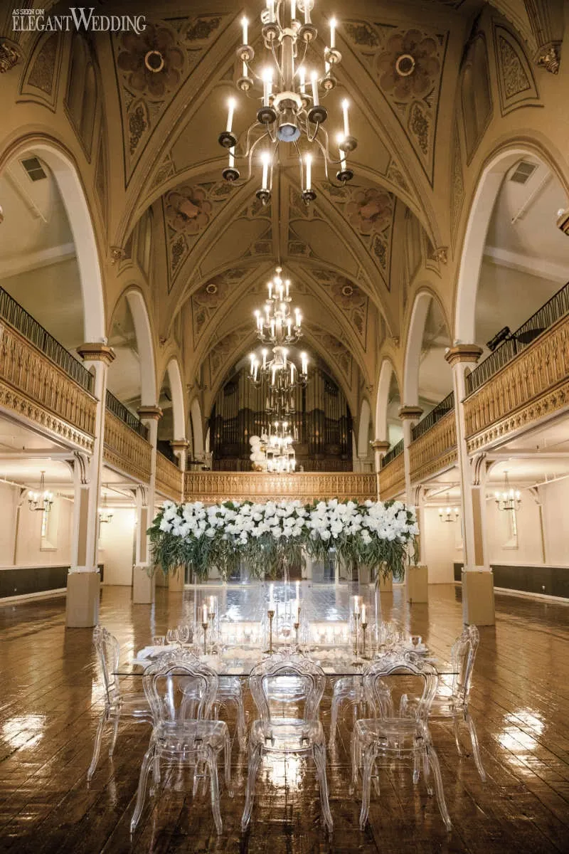 The image showcases Le Salon Richmond, a premier wedding venue listed under Mariage Elegant, Canada. It features an opulent, arched hall with intricately painted ceilings and elegant chandeliers casting a warm glow over the space. In the center, a grand display of white flowers is arranged on a long, reflective table surrounded by transparent, modern chairs. The polished wooden floor adds a rich, inviting texture, making this venue an ideal setting for a sophisticated and memorable wedding celebration.