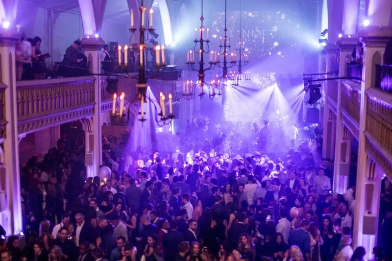 The image shows a bustling wedding reception at Le Salon Richmond, a premier wedding venue in Canada. The venue features elegant, arched ceilings adorned with numerous chandeliers casting a warm glow over a lively crowd. Guests are seen mingling and dancing under vibrant purple lighting, creating a festive and joyful atmosphere. The spacious area is perfectly suited for large gatherings, highlighting the venue's capacity and suitability for grand wedding celebrations.