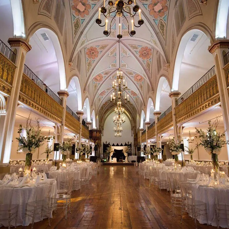 The image features a stunning wedding venue, Le Salon Richmond, categorized under Wedding Venues on Mariage Elegant, Canada. The venue showcases a grand, elegantly decorated banquet hall with high arched ceilings adorned with intricate floral designs and ornate chandeliers casting a warm glow. The space is arranged with multiple round tables, each elegantly set with white linens, clear glassware, and flower centerpieces, creating an inviting atmosphere. The wooden flooring enhances the venue's classic and sophisticated ambiance, perfect for hosting luxurious wedding celebrations.