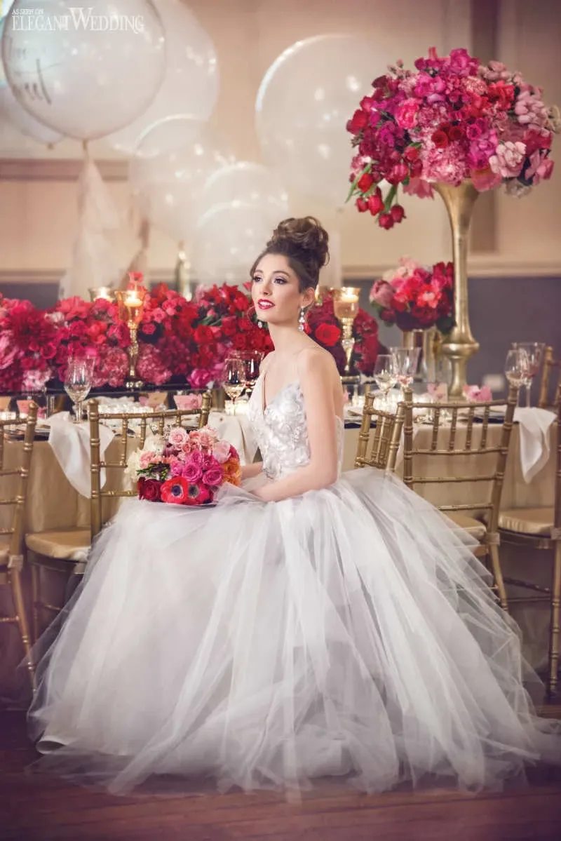 The image showcases a beautifully decorated wedding venue by Le Salon Richmond, categorized under Wedding Venues. A bride in a stunning white ballgown sits gracefully at a lavishly set table adorned with vibrant red and pink floral arrangements. The setting includes elegant gold chairs and delicate candles that enhance the romantic atmosphere. Large white balloons in the background add a whimsical touch, making it an ideal setting for an elegant wedding celebration.