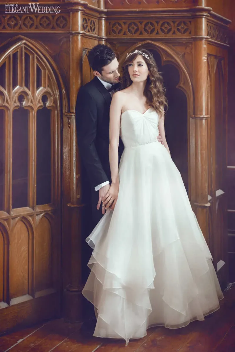 A bride and groom in elegant wedding attire stand posed against a richly detailed wooden backdrop with gothic-style arches, part of the interior at Le Salon Richmond, a renowned wedding venue in Canada. The bride wears a strapless, layered white gown and a delicate headpiece, while the groom is dressed in a classic black suit. This sophisticated setting highlights the venue's timeless and romantic ambiance, perfect for a wedding celebration.