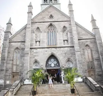 The image shows an elegant stone church with a tall, pointed tower, serving as the wedding venue Le Salon Richmond, which falls under the Wedding Venues category for Mariage Elegant in Canada. The church's grand entrance is adorned with large potted plants on either side, creating a welcoming atmosphere for guests. A few people are seen ascending the wide staircase leading to the entrance, enhancing the sense of anticipation and celebration common at wedding events. The overall impression is one of historic charm and sophistication, ideal for a memorable wedding ceremony.