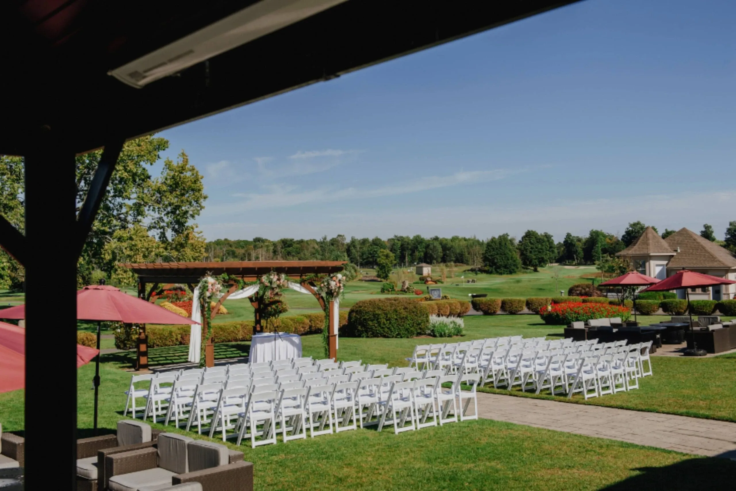 The image shows a beautifully arranged outdoor wedding venue at Golf Saint-Raphaël, categorized under Wedding Venues. Rows of white chairs are neatly set on a lush green lawn, facing a wooden arch adorned with floral decorations, perfect for a wedding ceremony. The background features manicured gardens and a clear blue sky, creating a serene and picturesque setting. This venue offers a charming and elegant atmosphere for couples looking to celebrate their special day in Canada.