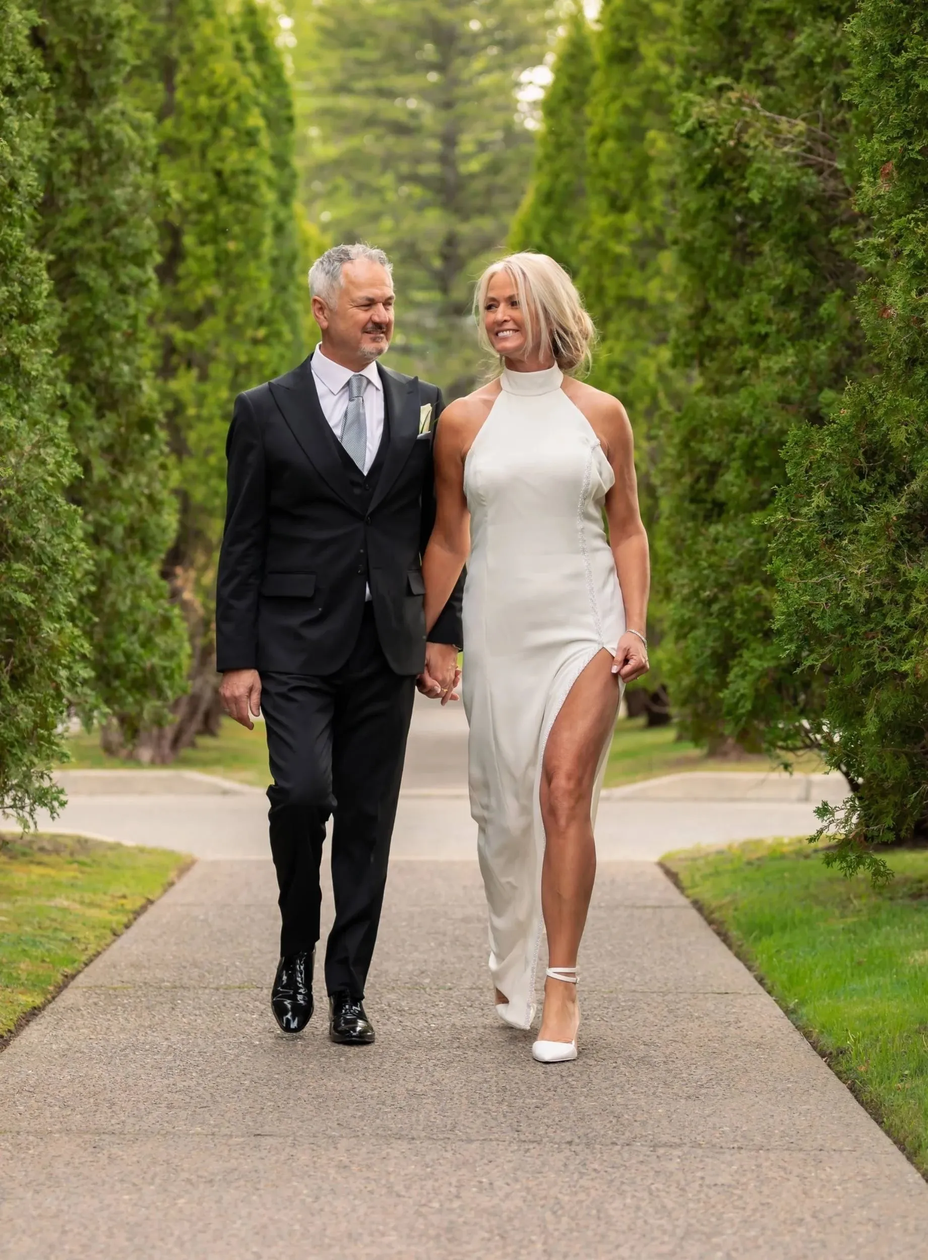 A couple, dressed elegantly, walks hand in hand down a scenic pathway at Golf Saint-Raphaël, a renowned wedding venue in Canada. The man is wearing a classic black suit with a white shirt and tie, while the woman is in a stylish white gown with a high neckline and a thigh-high slit. They are surrounded by lush green trees, creating a romantic and serene atmosphere perfect for weddings. The setting exemplifies the charm and elegance typical of Golf Saint-Raphaël as a top choice for wedding celebrations.