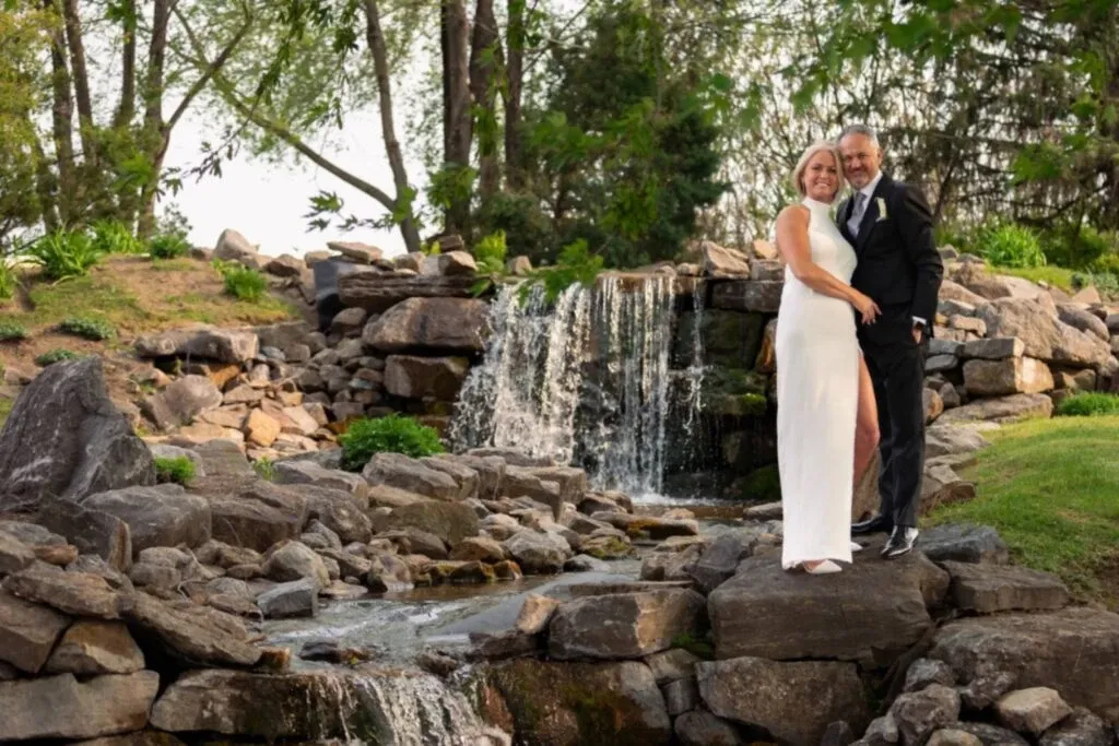 The image features a couple standing beside a picturesque waterfall at Golf Saint-Raphaël, a wedding venue in Canada. The bride is wearing a sleek, white gown, while the groom is dressed in a classic black suit, exuding elegance. They are surrounded by natural greenery and artfully arranged rocks, creating a serene and romantic backdrop ideal for weddings. The scene captures the essence of a tranquil outdoor wedding venue, perfect for creating memorable moments.