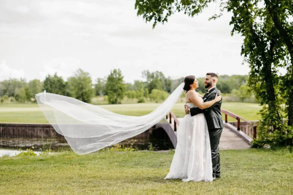 A happy couple embraces on their wedding day at Golf Saint-Raphaël, a stunning wedding venue. The bride, wearing an elegant lace gown with a long flowing veil, stands beside the groom, dressed in a sharp black suit. They are positioned in front of a picturesque wooden bridge surrounded by lush greenery. The expansive golf course in the background enhances the serene and romantic atmosphere, making it an ideal location for wedding celebrations.