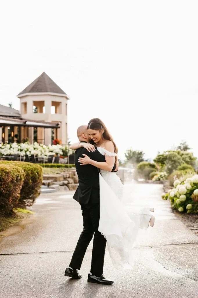 A newlywed couple shares a joyful embrace outside Golf Saint-Raphaël, a premier wedding venue in Canada. The groom, dressed in a classic black suit, lifts the bride, who is wearing an elegant white gown. They are set against a picturesque backdrop of manicured gardens and a charming building with a turret, adding to the romantic ambiance of this celebrated wedding location. The pathway is lined with vibrant greenery and blooming flowers, enhancing the serene and beautiful setting at this exquisite venue.