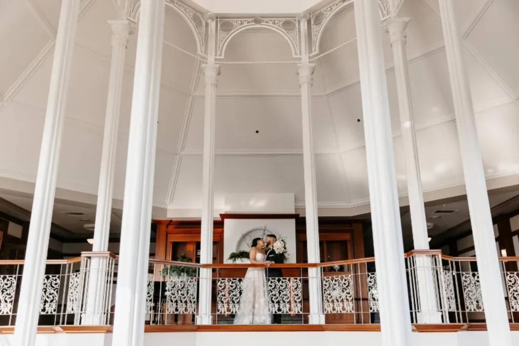 The image shows a beautifully decorated wedding venue at Golf Saint-Raphaël, part of Mariage Elegant's wedding venue listings in Canada. A bride stands elegantly on a balcony, surrounded by intricate white railings and tall, white pillars that lead up to a high, vaulted ceiling. The ornate architecture creates a sense of grandeur and sophistication, ideal for an unforgettable wedding ceremony. The bride is holding a bouquet of flowers, enhancing the romantic atmosphere of the scene. The venue’s elegant interior design makes it a perfect choice for couples seeking a classic and stylish wedding location.