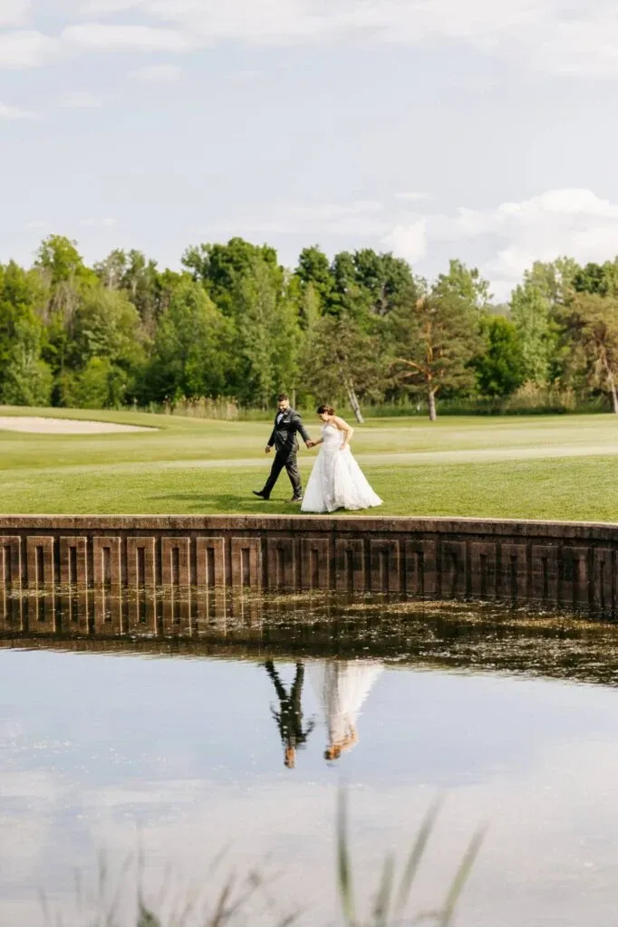 A bride and groom are walking hand in hand along a lush, green golf course at Golf Saint-Raphaël. The serene landscape, perfect for wedding ceremonies and receptions, features a wooden barrier bordering a calm pond, reflecting the couple's image in the water. This picturesque setting is ideal for creating unforgettable wedding memories, making it a prime choice for those seeking elegant wedding venues.