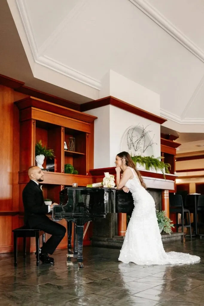 A couple is engaged in a tender moment at Golf Saint-Raphaël, a wedding venue in Canada. The bride, wearing an elegant white gown, leans thoughtfully against a grand piano, while the groom, dressed in a classic black suit, sits at the piano, looking upwards at her. The setting is sophisticated, with wooden paneling and lush green plants adorning the space, creating a warm and inviting atmosphere ideal for weddings. The piano area adds a touch of elegance and serves as a perfect backdrop for capturing memorable wedding moments.