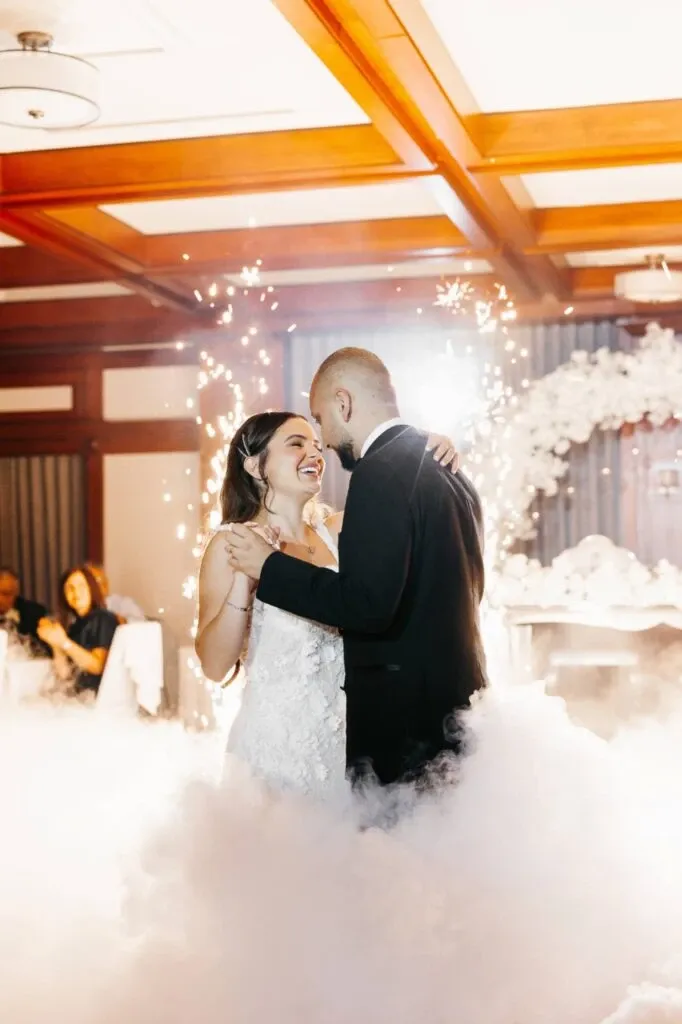 A bride and groom share their first dance amidst a cloud of romantic mist and vibrant sparklers at Golf Saint-Raphaël, a premier wedding venue in Canada. The joy on their faces reflects the perfect ambiance created by the elegant setting. The room features a beautifully decorated arch adorned with flowers in the background, adding to the overall charm of the venue. This image captures a memorable wedding moment, showcasing the venue's ability to provide a magical and elegant setting for such special occasions.