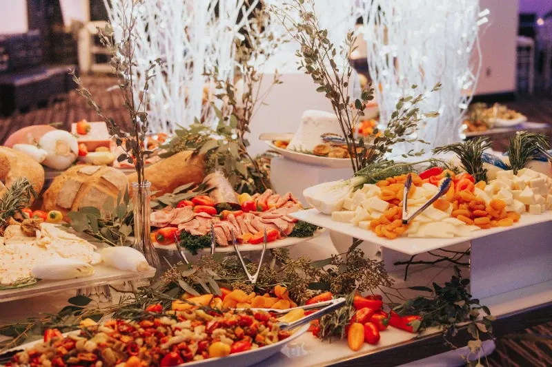 The image showcases an elegant wedding buffet at Plaza Centre Ville, a premier wedding venue in Canada, under the category of Wedding Venues on Mariage Elegant. The buffet table is adorned with an assortment of gourmet cheeses, charcuterie, and fresh bread. Delicate garnishes and rustic greenery enhance the presentation, creating a sophisticated and inviting atmosphere for wedding guests. The lighting accentuates the vibrancy of the fresh produce and intricate design of the food arrangements, adding to the venue's refined ambiance.