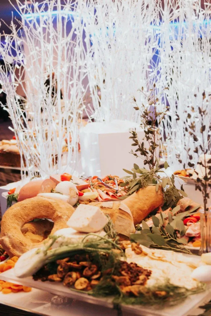 The image showcases an elegant wedding setting at Plaza Centre Ville, a renowned wedding venue featured on Mariage Elegant, Canada. The focal point is a beautifully arranged buffet table adorned with an assortment of gourmet cheeses, savory pastries, and fresh herbs, artfully displayed on a white platter. In the background, delicate white decorative branches add a sophisticated and ethereal ambiance, enhancing the venue's luxurious atmosphere. The overall presentation captures the essence of a high-class wedding reception, emphasizing the attention to detail and quality offered by this venue.