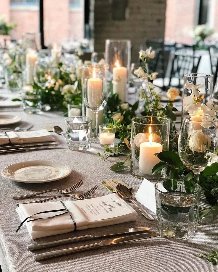 The image showcases an elegantly set wedding reception table at Plaza Centre Ville, a wedding venue featured on Mariage Elegant, Canada. The table is beautifully arranged with white and cream floral centerpieces interspersed with lush greenery, creating a romantic ambiance. Tall and short glass candle holders with lit candles add a warm, inviting glow to the setting. Each place setting includes polished cutlery, glassware for water and wine, crisp white napkins tied with a black ribbon, and personalized menu cards, emphasizing attention to detail. The soft lighting and tasteful decor highlight the sophisticated atmosphere of this wedding venue.