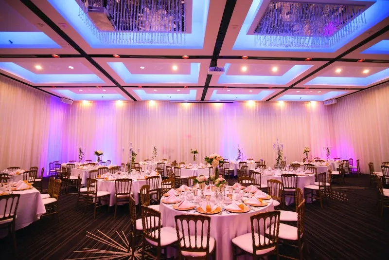 The image showcases a beautifully decorated wedding venue at Plaza Centre Ville, categorized under Wedding Venues for Mariage Elegant in Canada. The spacious room features elegantly arranged round tables adorned with white tablecloths, fine china, and delicate floral centerpieces. The ambient lighting casts a warm, inviting glow with soft pink and purple hues illuminating the room. The ceiling is adorned with intricate chandeliers that add a touch of sophistication to the venue. Wooden chairs with plush cushions encircle the tables, creating an atmosphere of comfort and elegance, perfect for an intimate wedding celebration.