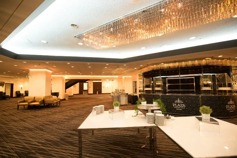The image shows the interior of Plaza Centre Ville, categorized under wedding venues, featuring a spacious and elegantly designed reception area. The room has modern decor with a large, glittering chandelier hanging from the ceiling, casting a warm glow over the space. The carpet has a contemporary pattern, and sleek white tables are arranged for a stylish appearance. Small greenery and decorative elements add a touch of elegance. The reception desk prominently displays the Plaza Centre Ville branding, contributing to the venue’s luxurious and sophisticated ambiance, ideal for hosting weddings and large events.