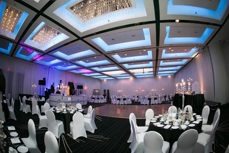 This image showcases the elegant wedding venue Plaza Centre Ville in Canada, under the wedding venue category. The spacious banquet hall is beautifully arranged for a wedding reception, featuring numerous round tables adorned with pristine white tablecloths and chairs draped in white covers. The ceiling is impressive, with intricate, modern lighting that casts a sophisticated blue hue across the room. Stunning chandeliers add a touch of luxury, while the dance floor is prepared for celebrations. Meticulously arranged table settings and lavish decor highlight the venue's attention to detail, making it an ideal choice for any couple's special day.
